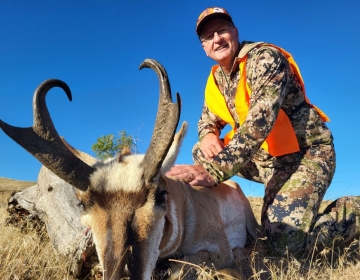 A smiling hunter, clad in camouflage and orange safety gear, kneels behind a pronghorn antelope with tall, curved horns in the bright Wyoming sunshine.